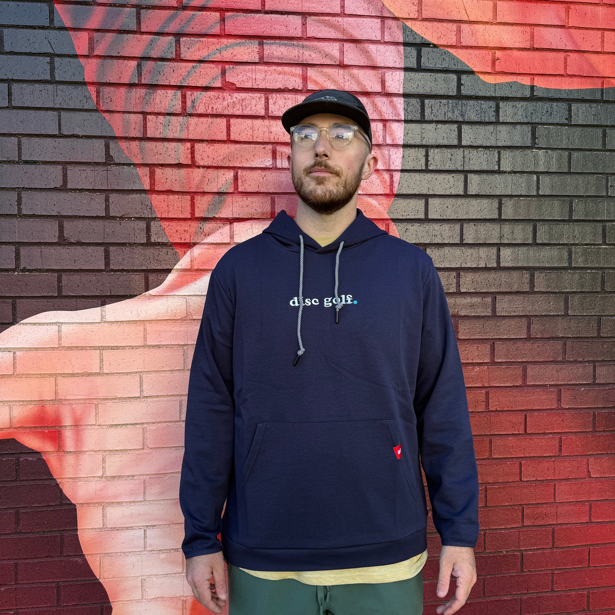 A man wearing a navy blue 'disc golf.' hoodie and green shorts stands in front of a brick wall with a red and black mural.